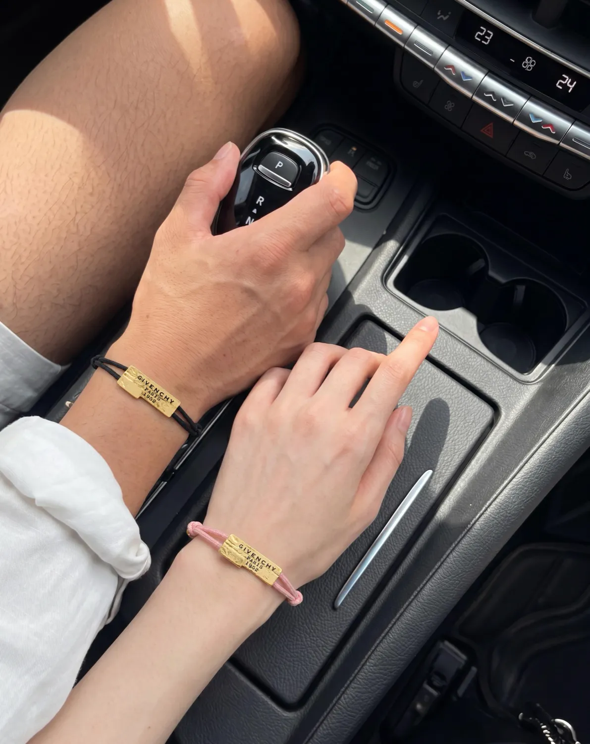 Two hands with gold and pink bracelets at a car's gear shift